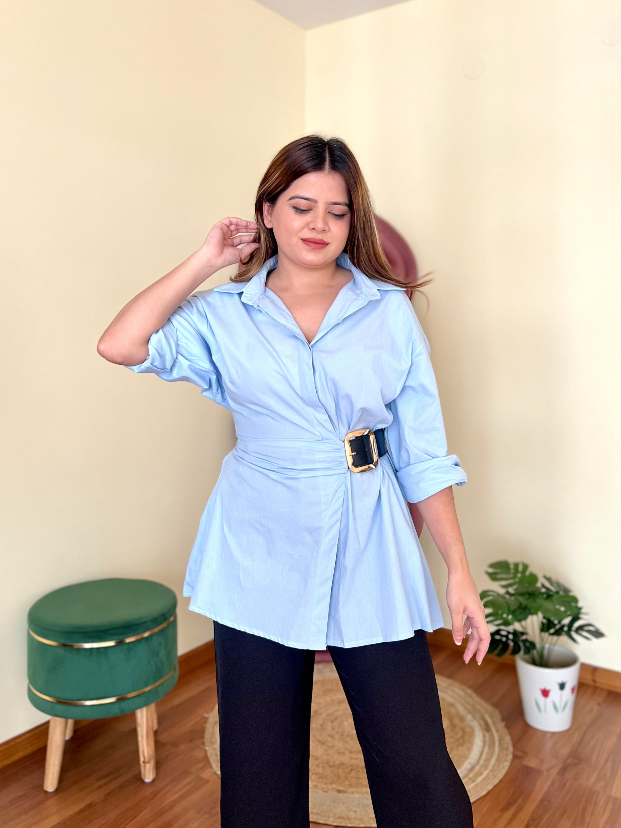 Woman wearing a light blue shirt with a belt in a room with a green stool and plant.